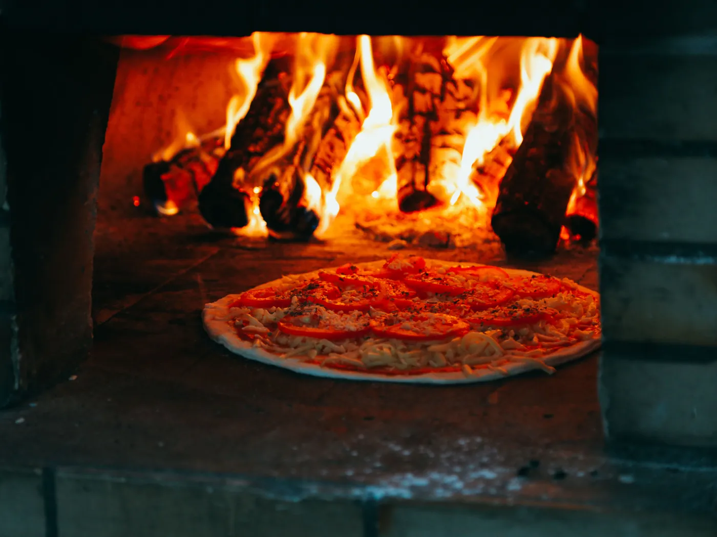 A tomato pizza on the floor of a brick oven beneath a roaring wood fire