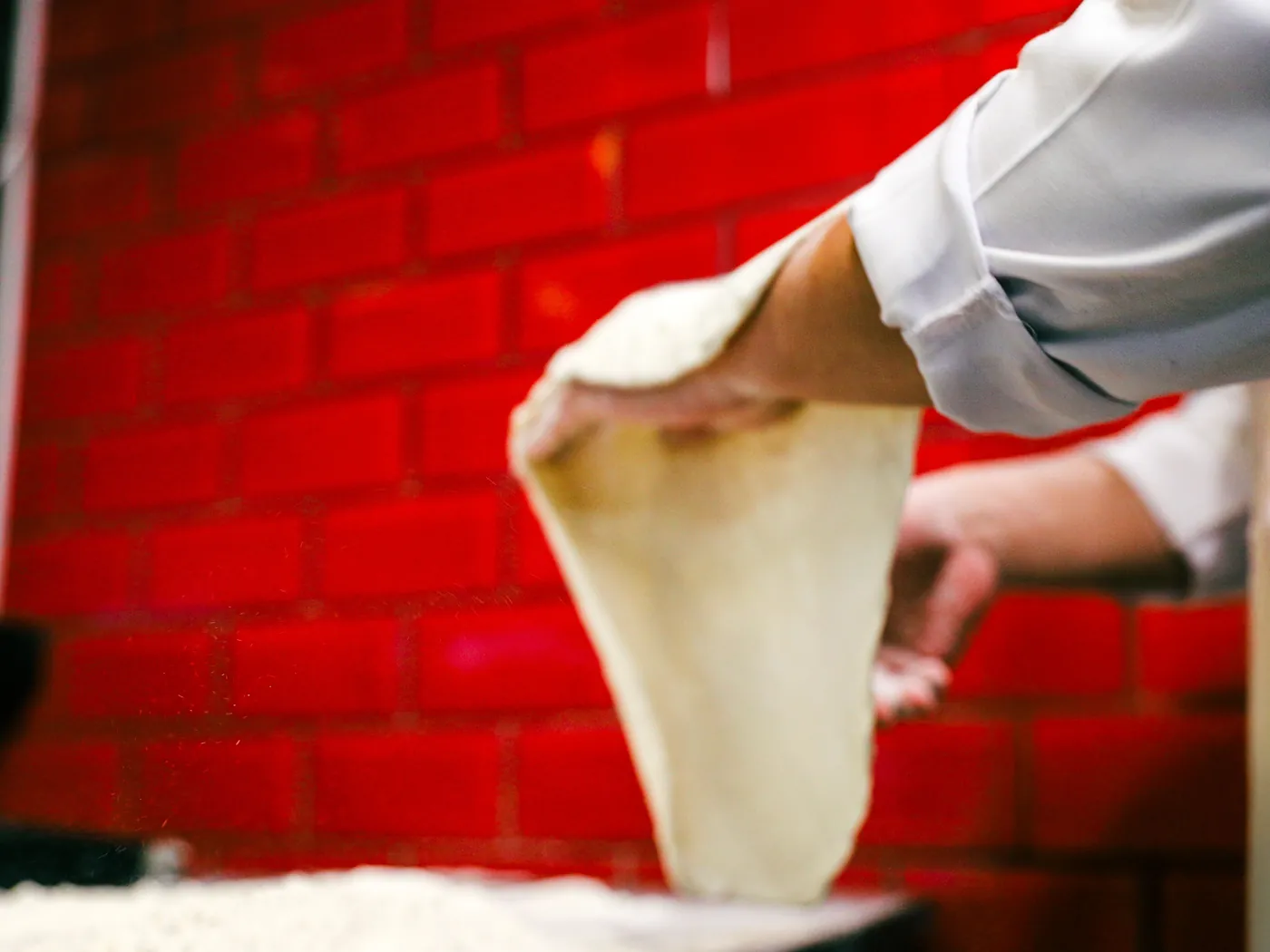 A cook in a white jacket lifting a stretched round of pizza dough in front of a red brick wall